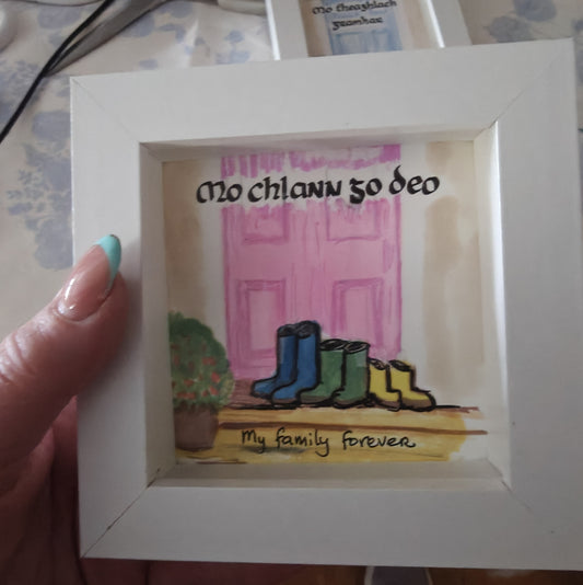 Handmade Irish family frame with three pairs of little wellies on a doorstep before a soft blush pink door. Hand-lettered Gaelic text 'Mo chlann go deo' meaning My family forever. Alcohol ink art in a white shadow box frame.