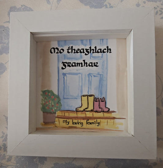 Handmade Irish family frame with two pairs of little wellies on a doorstep before a pale duck-egg blue door. Hand-lettered Gaelic text 'Mo Teaghlach Grámhar' meaning My loving family. Alcohol ink art in a white shadow box frame.